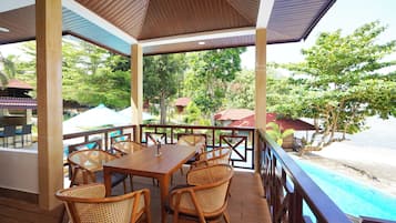 Un espace extérieur aménagé avec des tables et des chaises en bois, protégé par un auvent, et offrant une vue sur une piscine et une plage.