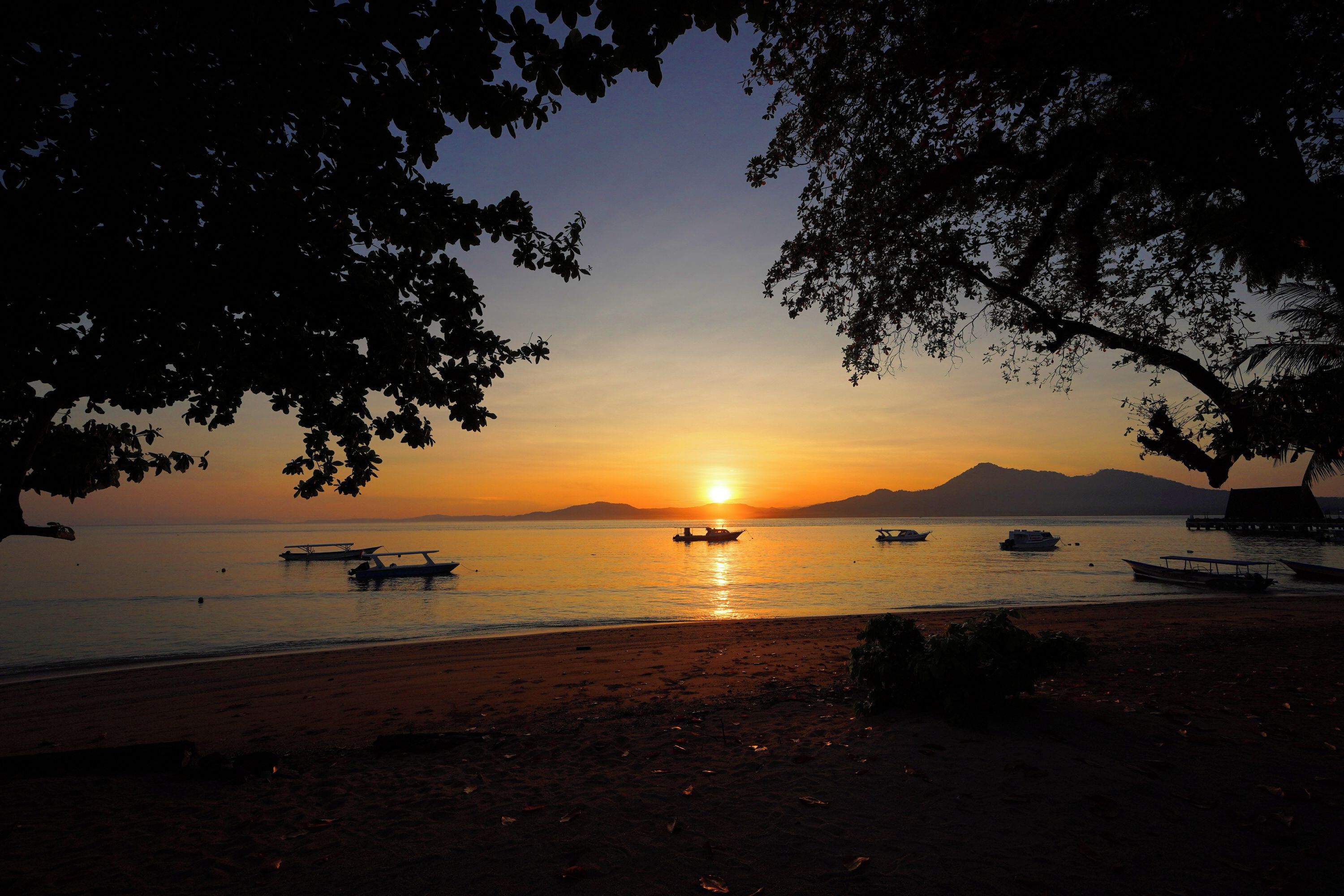 Une scène de plage avec des bateaux, un coucher de soleil et des montagnes en arrière-plan.