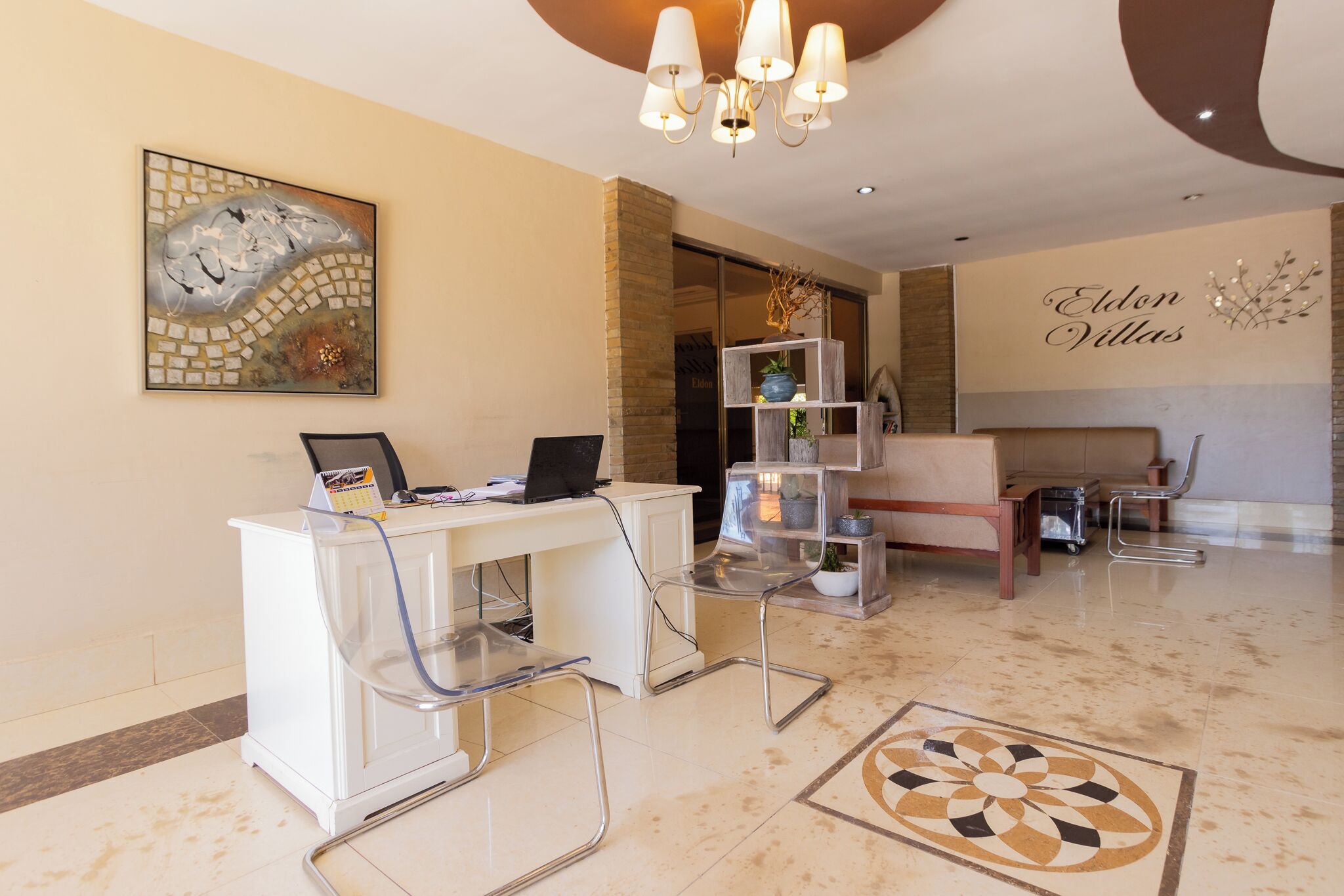 Ein moderner Empfangsbereich mit Schreibtisch, Stühlen, einer Chandelierleuchte, einem Wandbild und einem Schild mit der Aufschrift 'Eldon Villas'.