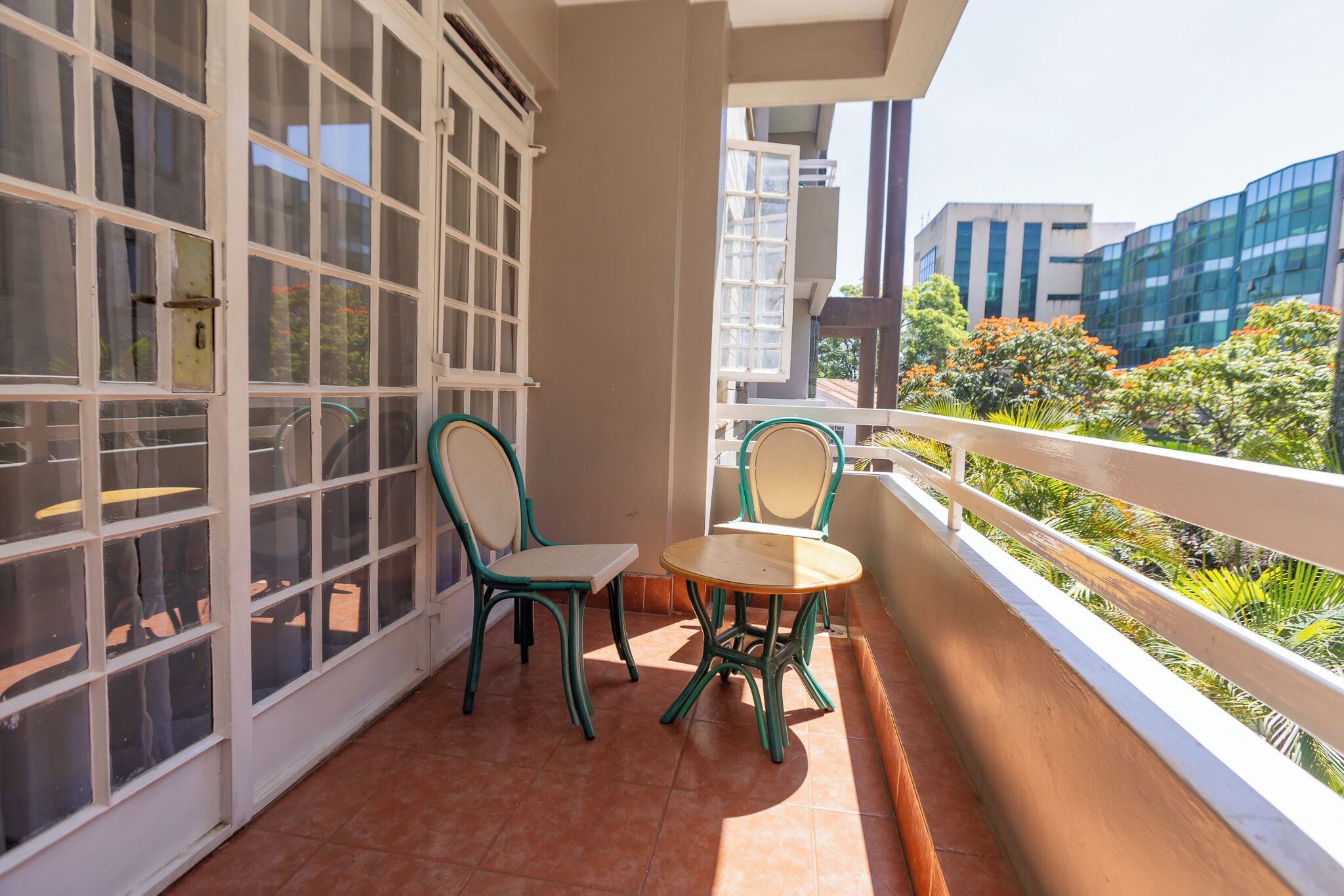 Ein Balkon mit Tisch und Stühlen, Blick auf Grünflächen und moderne Gebäude, sowie ein großes Fenster mit mehreren Fenstern.