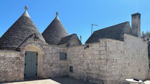 A stone building with a central door, two conical roofs, and a small table with a chair. - Trullo Cinquenoci Jr Suite (Locorotondo)