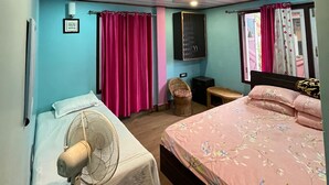 A bedroom with a bed, a chair, a small table, a fan, and a window with curtains. - Manjushree Homestay (Darjeeling)