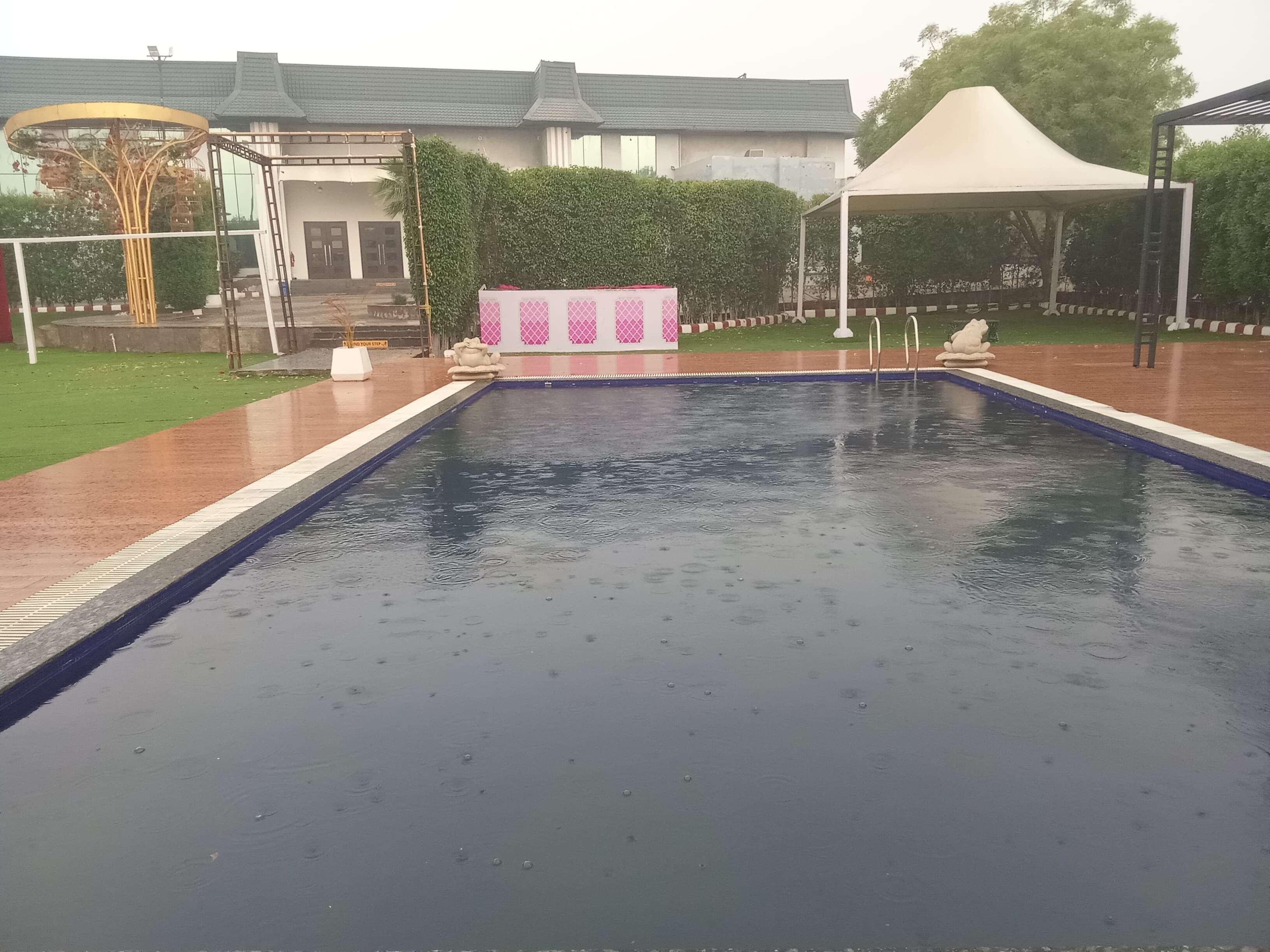A swimming pool with a surrounding deck, a covered seating area, and a building in the background.