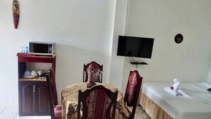 Une petite pièce comprenant une table et des chaises, un téléviseur fixé au mur, un lit avec un matelas blanc et un petit meuble contenant des ustensiles de cuisine.