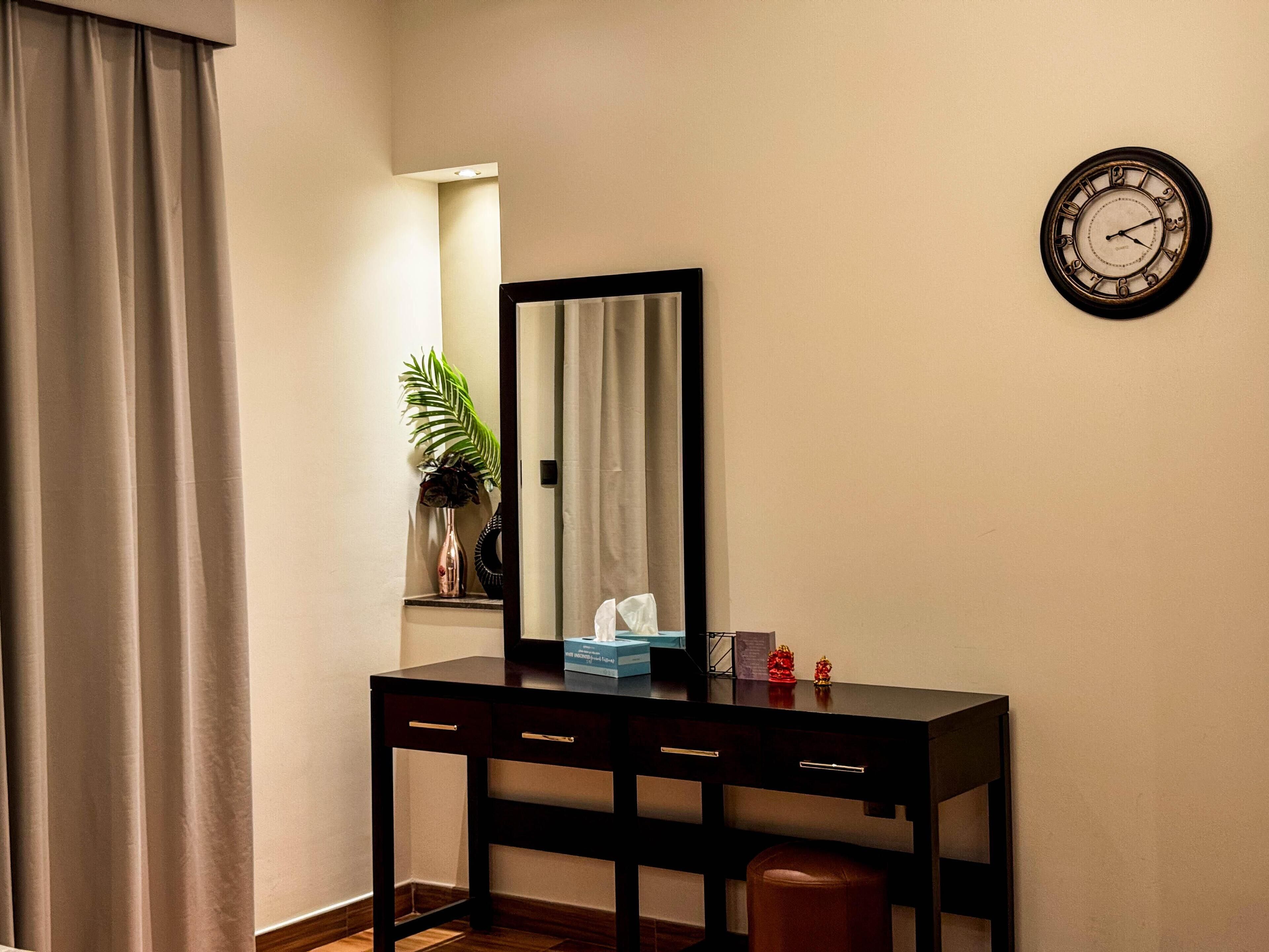 A room with a black desk, a mirror, a clock, a plant, and a curtain.
