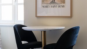 Un coin repas avec une table ronde et deux chaises noires, une affiche encadrée au mur et une fenêtre avec des stores.