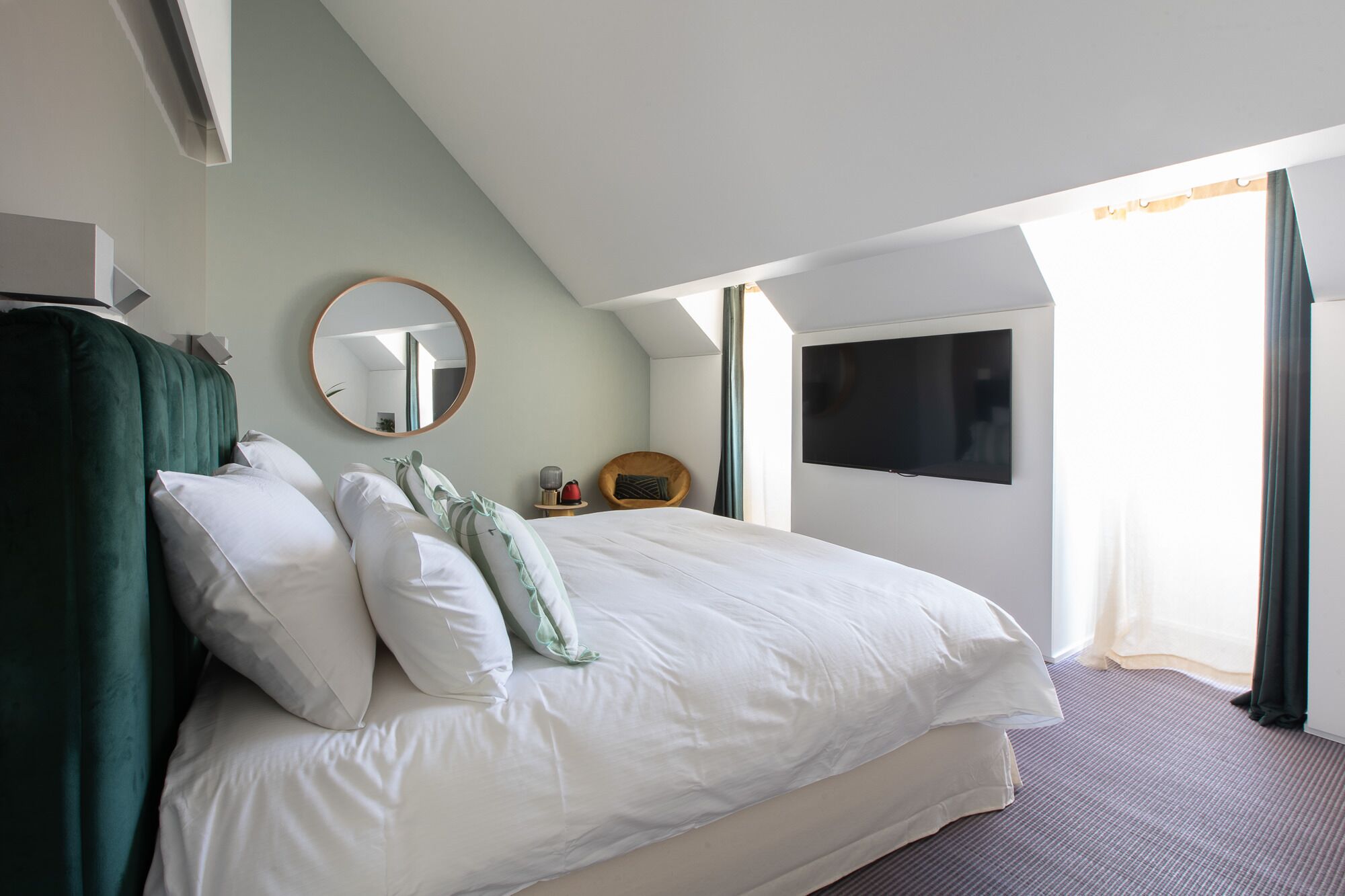 A modern hotel room with a large bed, a round mirror, a television, and a window with curtains.