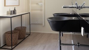 Une salle de bain moderne avec un grand miroir, deux lavabos noirs et un porte-serviettes. Un tableau encadré est accroché au mur, et une petite table avec des paniers se trouve dans la pièce.