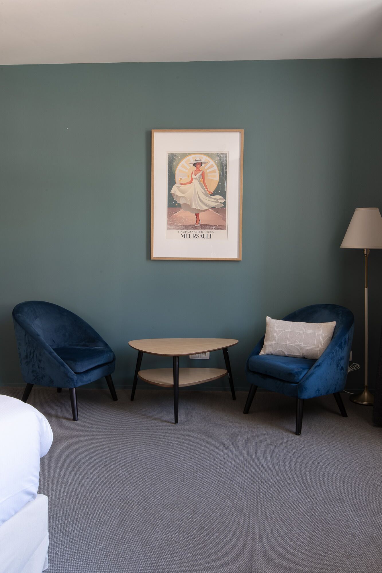 Una habitación con dos sillones tapizados en terciopelo azul, una mesita redonda y un cartel enmarcado en la pared.