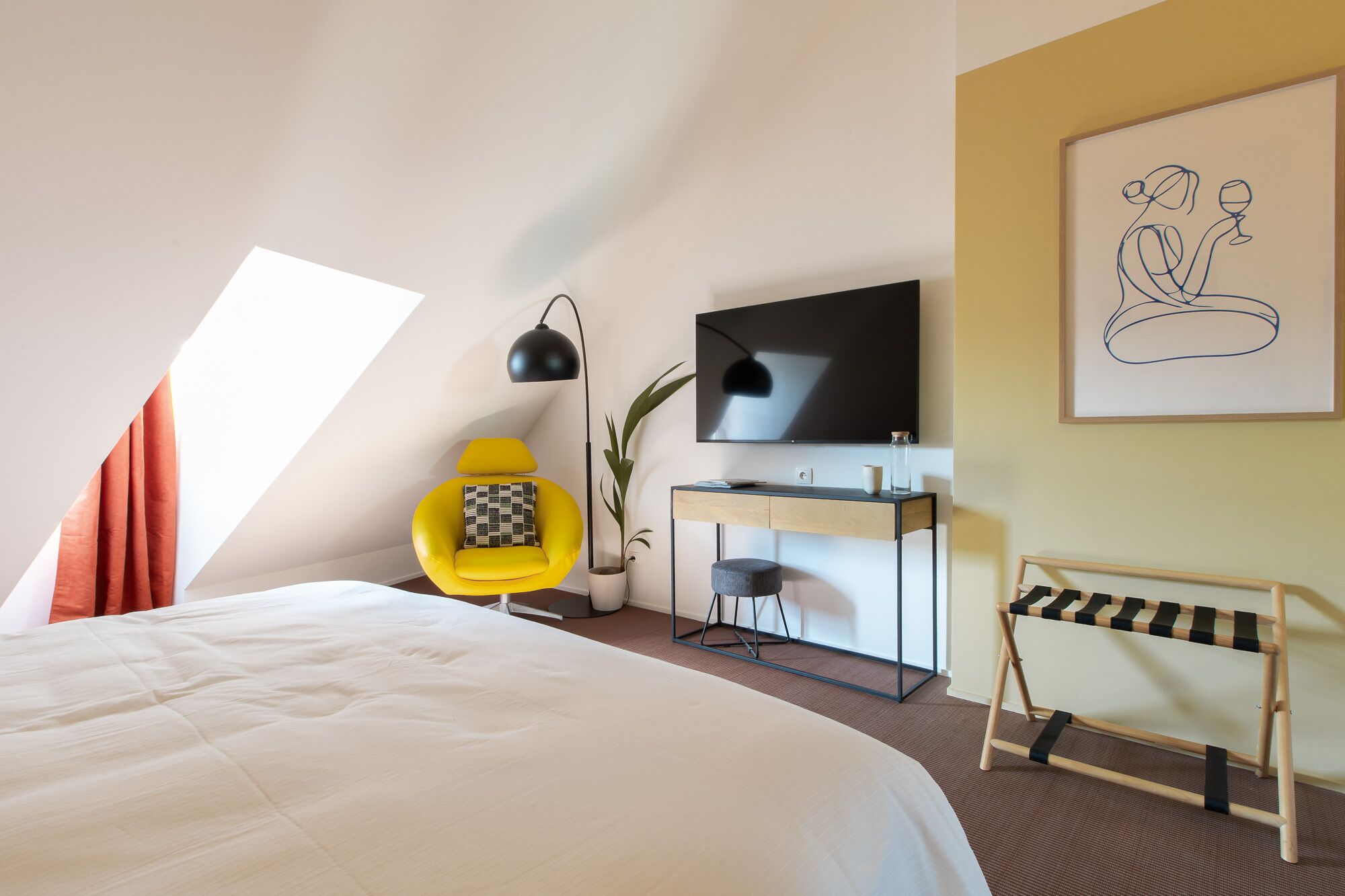 A modern hotel room with a large bed, a desk with a flat-screen TV, a yellow armchair, and a painting on the wall.