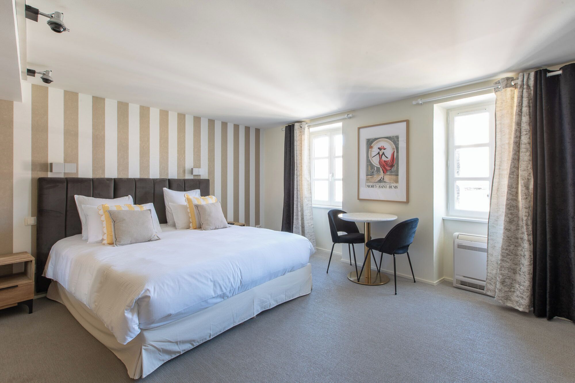 A hotel room with a large bed, a small table with two chairs, a framed picture on the wall, and a window with curtains.
