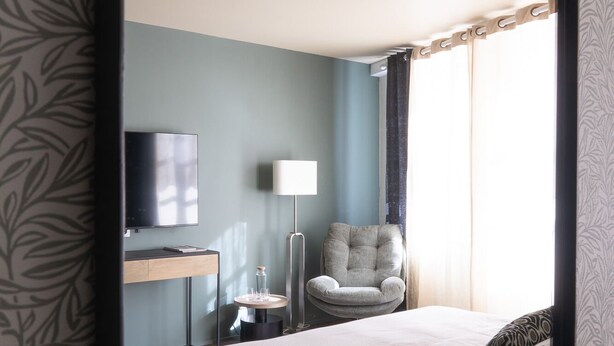 Une chambre d’hôtel comprenant un lit, un bureau avec un téléviseur à écran plat, une chaise, une lampe et une fenêtre avec des rideaux.