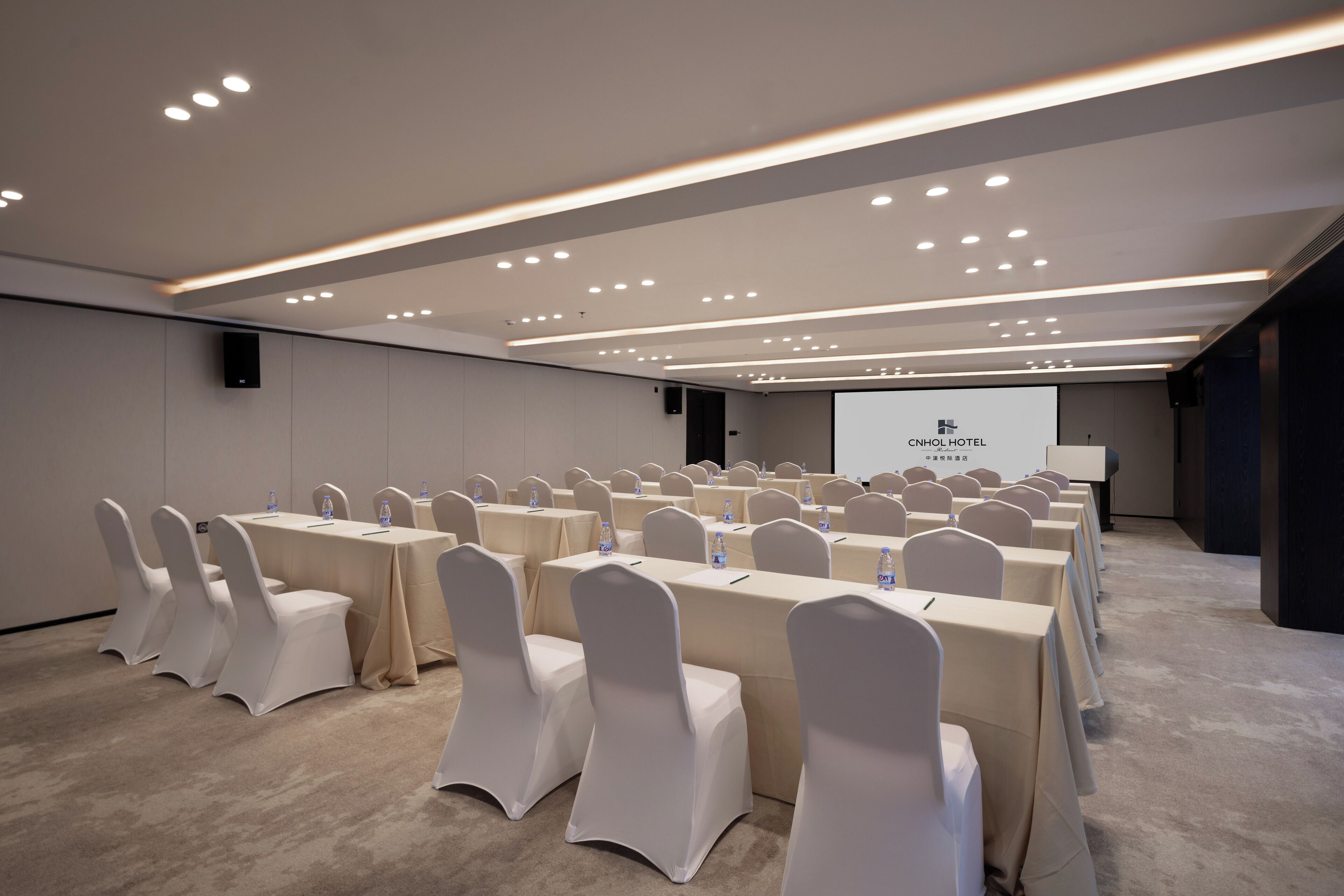 A conference room with rows of tables and chairs, a projector screen, and a speaker.