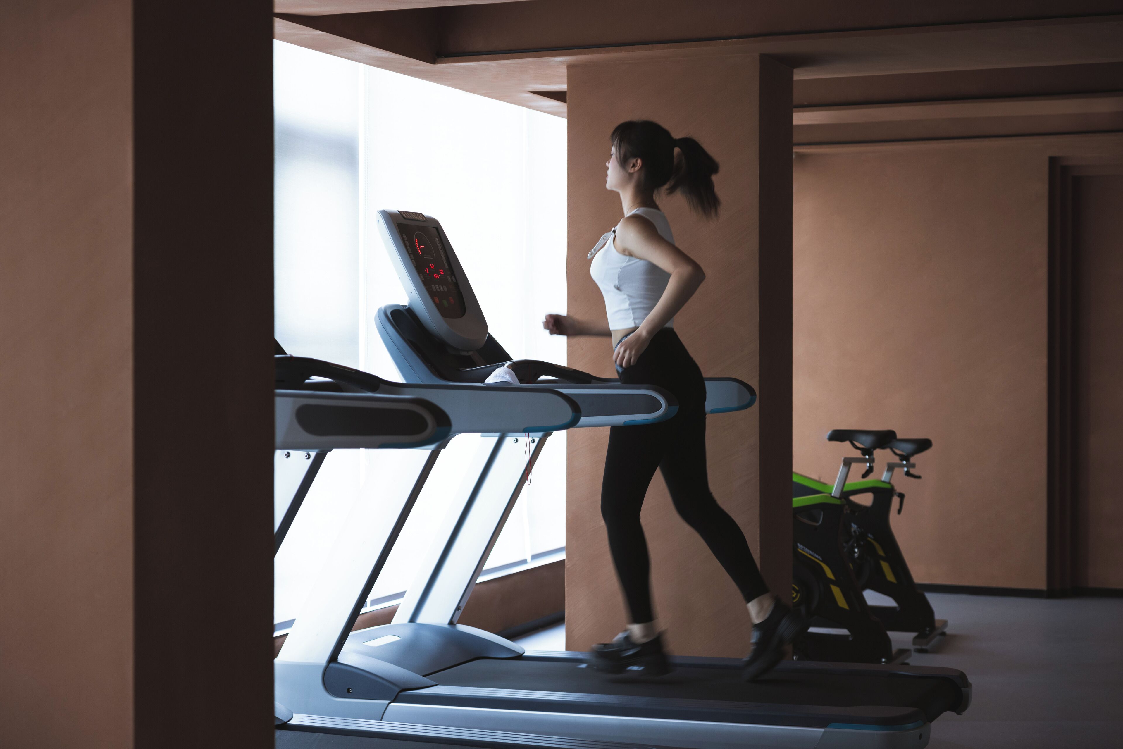 Eine Frau läuft auf einem Laufband in einem Fitnessstudio, im Hintergrund sind Trainingsfahrräder zu sehen.