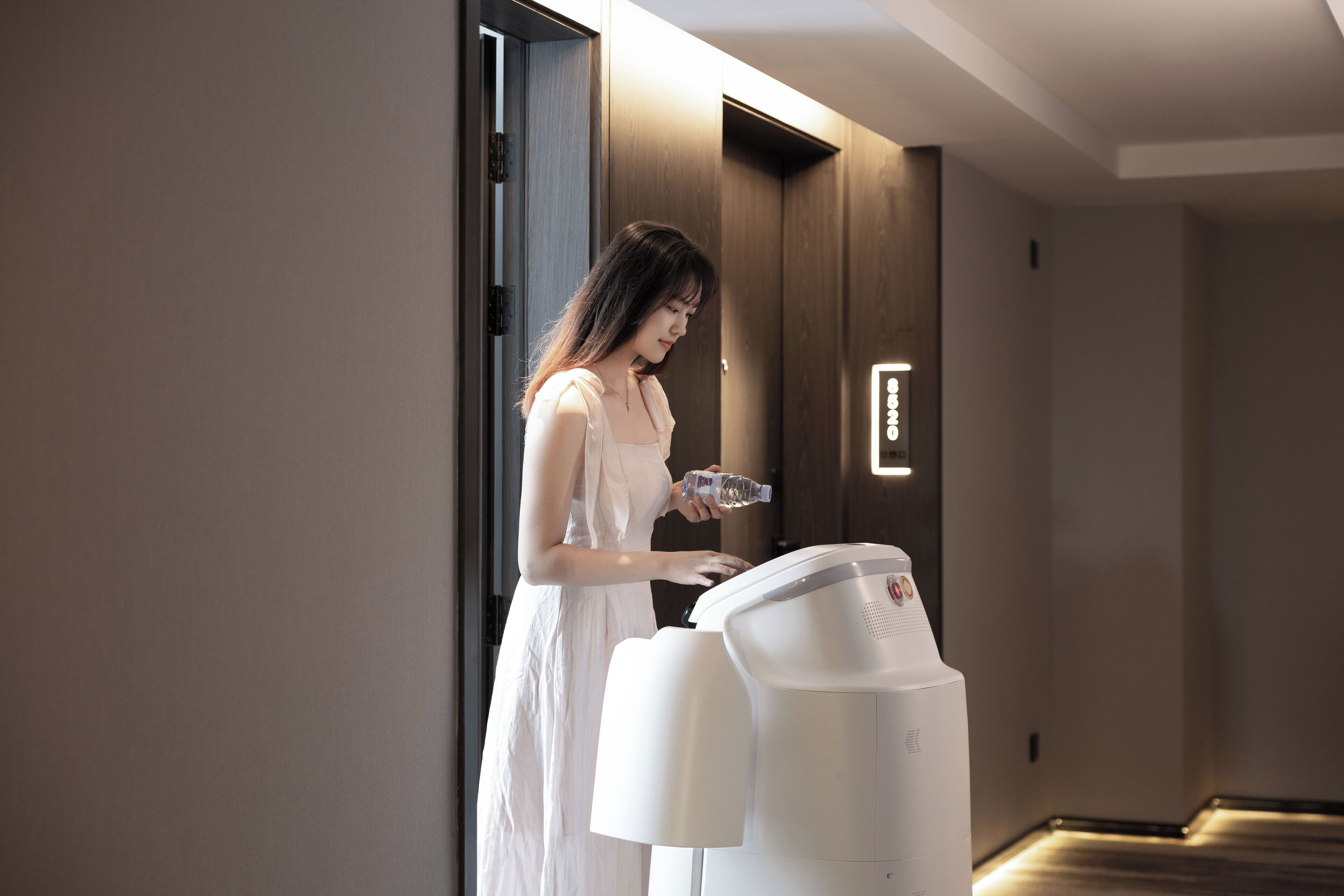 Eine Frau im weißen Kleid steht neben einem weißen, modernen Wasserspender in einer Halle mit Holzwänden und einer Tür.