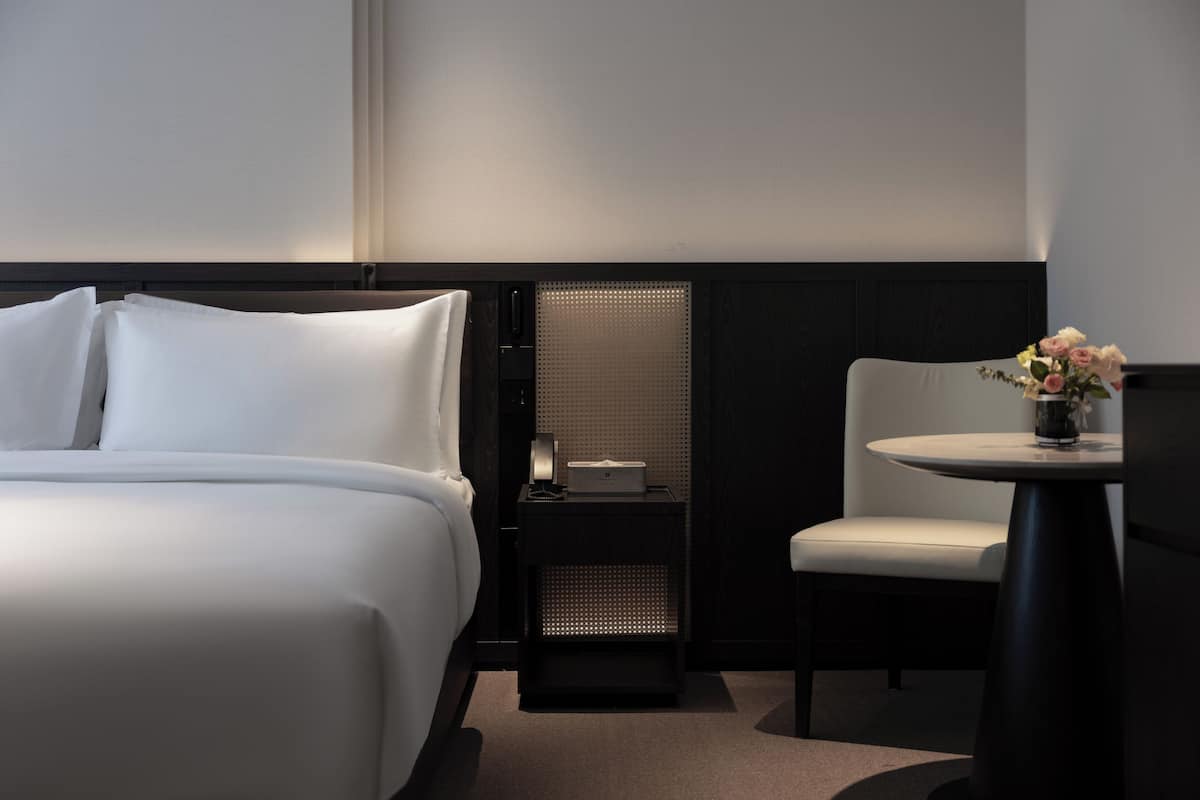 A hotel room with a neatly made bed, a small table with a vase of flowers, and a chair.
