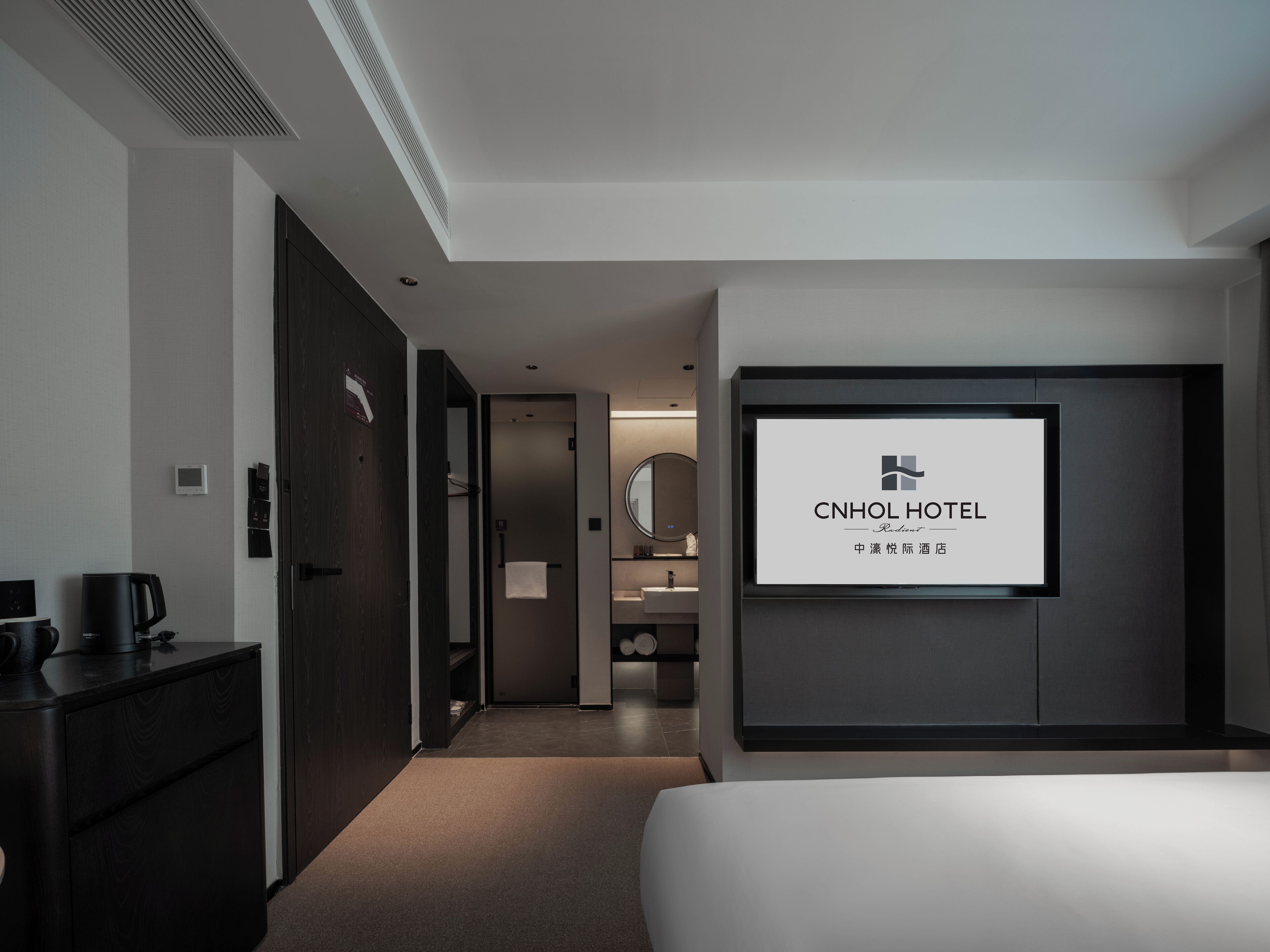 A modern hotel room with a large bed, a TV mounted on the wall, a desk with a coffee maker, and a wardrobe.