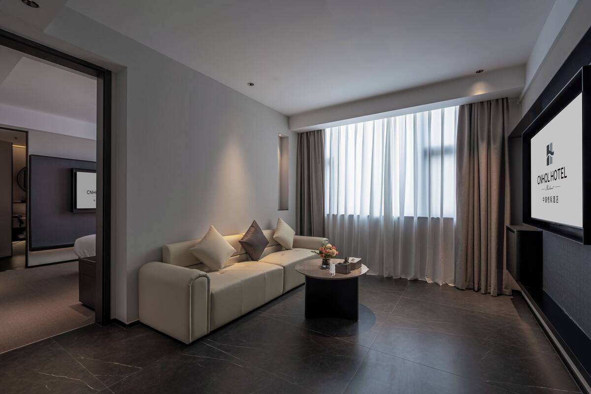 A modern hotel room with a large window, a sofa, a coffee table, and a TV screen displaying the hotel name.