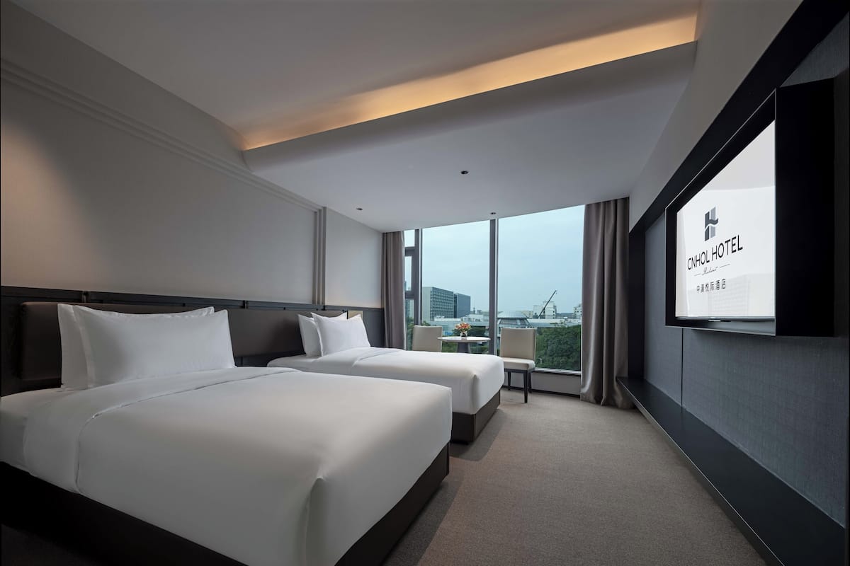 A hotel room with two beds, a TV, and a large window offering a city view.
