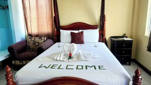 A hotel room with a bed featuring a 'WELCOME' sign, a wooden headboard, a nightstand, a chair, and a window with curtains. - See Belize Tranquil Sea View Studio With (Belize City)