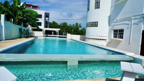 A rectangular swimming pool with a white deck, surrounded by white walls and a covered seating area.