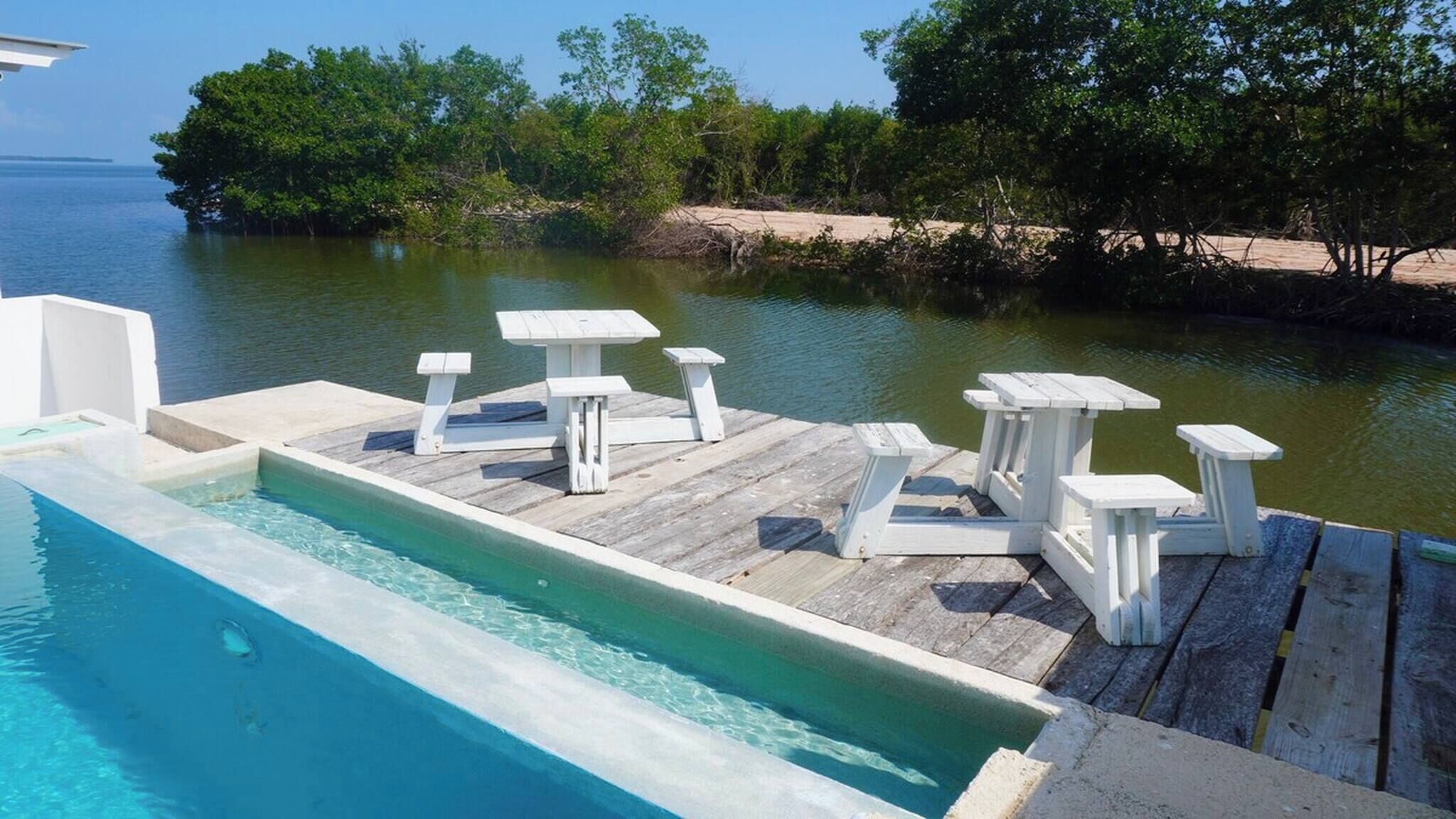 A wooden deck with a pool, white outdoor furniture, and a view of the water and trees.