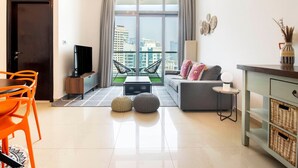 Ein modernes Wohnzimmer mit einer Couch, einem Couchtisch und einem Fernseher. Es gibt einen Essbereich mit orangefarbenen Stühlen und einem Tisch. Das Zimmer hat große Fenster mit Blick auf die Gebäude draußen.