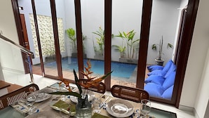 A dining area with a table set for four, large windows, and a view of a pool and outdoor seating. - Nusa Dua 3BR Villa • Perfect for Families (Bali)