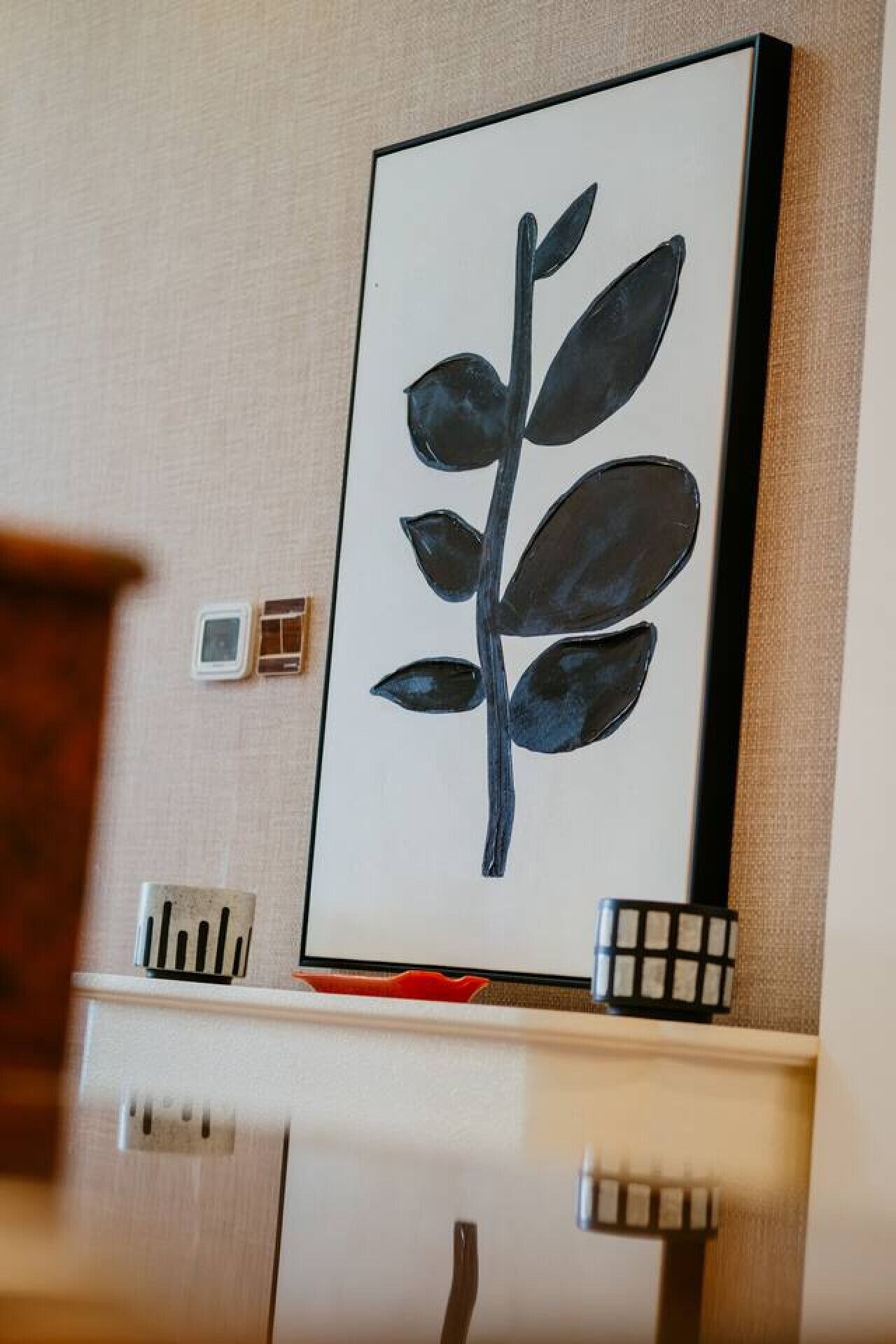 Une œuvre d’art encadrée représentant une feuille accrochée au mur, une petite horloge et un objet rouge posé sur une étagère.