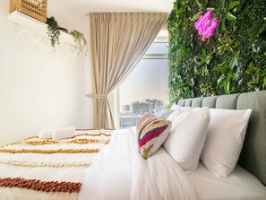 A modern bedroom with a bed, white bedding, a colorful pillow, a green wall with plants, and a city view.