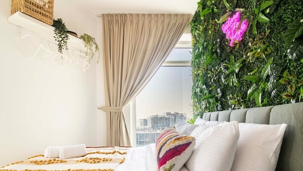 A modern bedroom with a bed, white bedding, a colorful pillow, a green wall with plants, and a city view. - Spacious 2BR in Downtown  | High Floor | 8 Guests (Dubai)