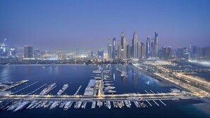 Un port de plaisance où de nombreux bateaux sont amarrés, une vue sur les grat-ciel d’une ville, et une étendue d’eau calme qui reflète les lumières de la ville.