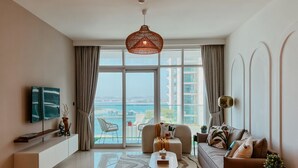 A modern living room with a large window, a sofa, a coffee table, and a TV on a wall. - Oceanfront Escape: Stylish 1 BR  in Beachfront (Dubai)