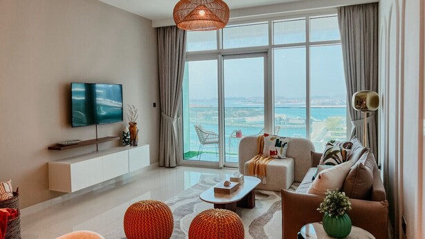 A modern living room with a large window, a sofa, a coffee table, and a TV mounted on the wall. - Oceanfront Escape: Stylish 1 BR  in Beachfront (Dubai)