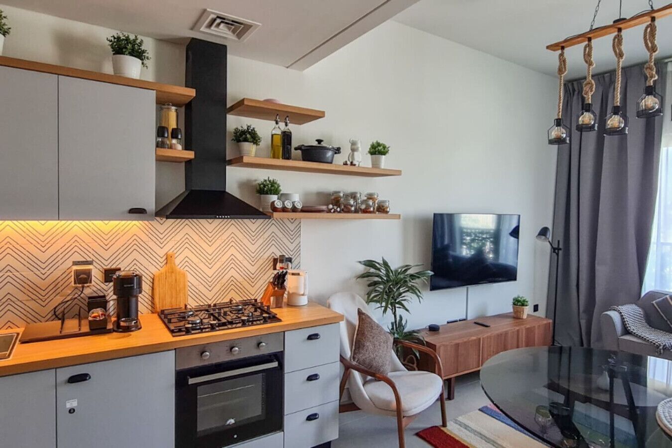 A modern kitchen with a gas stove, oven, and built-in cabinets. There is a dining area with a glass table and a sofa. A TV is mounted on the wall, and there are shelves with plants and kitchen items.