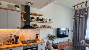 A modern kitchen with a gas stove, oven, and built-in cabinets. There is a dining area with a glass table and a sofa. A TV is mounted on the wall, and there are shelves with plants and kitchen items.