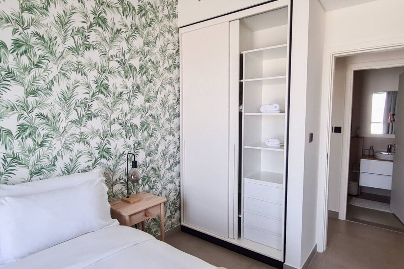 Une chambre à coucher comprenant un lit, une table de chevet, une armoire et une salle de bain visible à travers une porte ouverte.