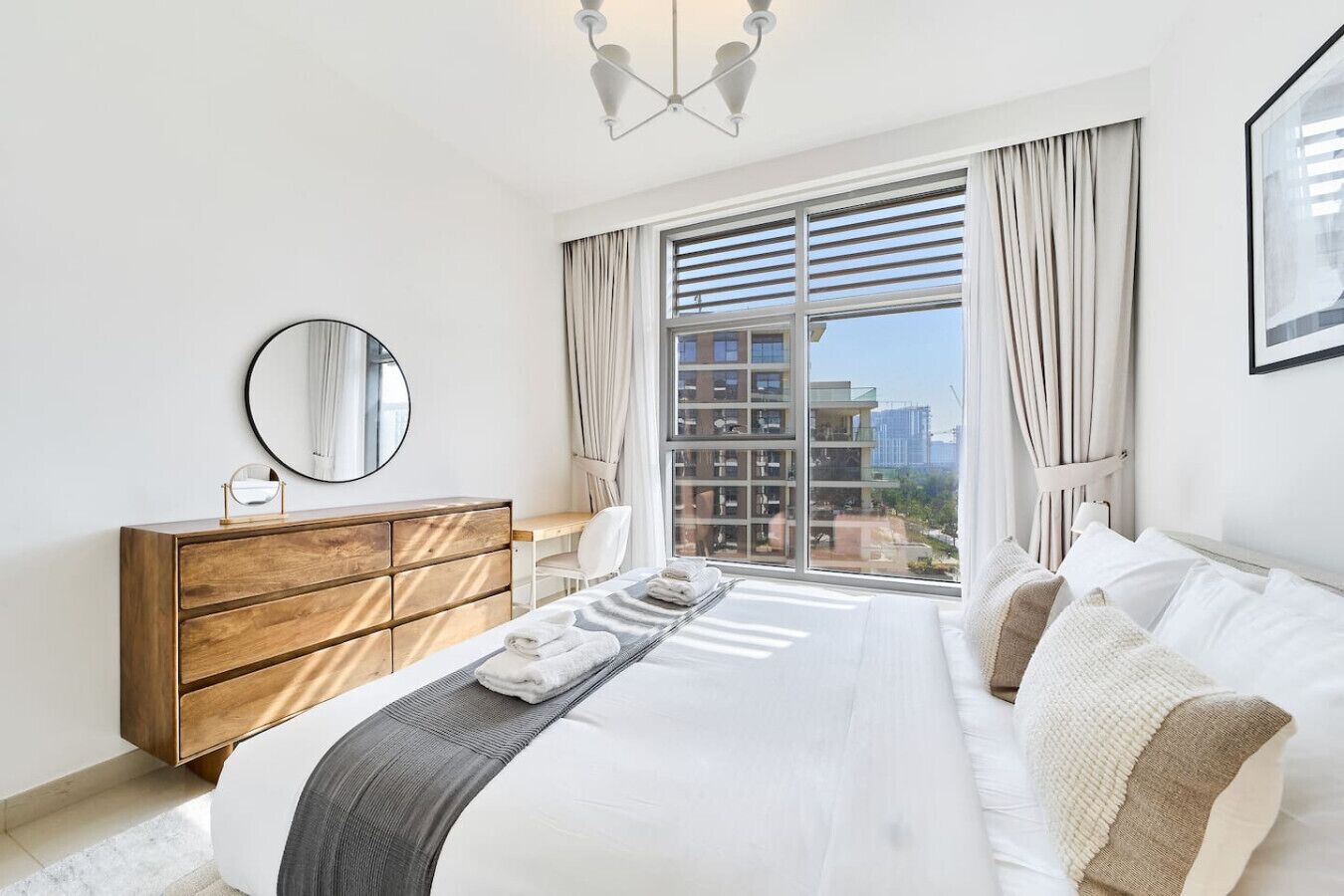 A modern hotel room with a large bed, a wooden dresser, a round mirror, and a view of a cityscape through floor-to-ceiling windows.