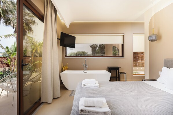 A modern hotel room with a large bed, a bathtub, a TV, and a balcony with plants.