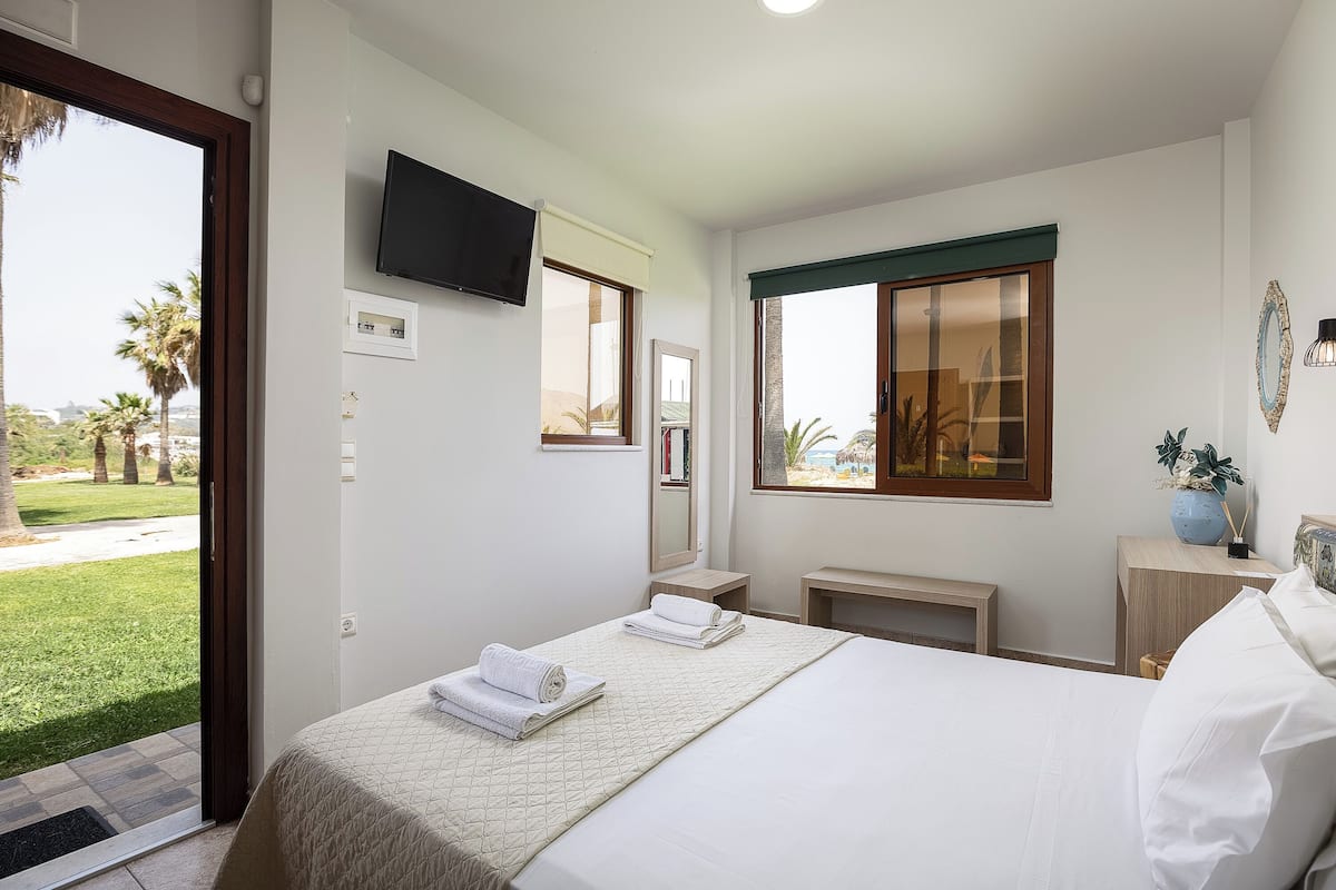 Una camera d'albergo con un letto, una televisione, una finestra vista palme e una porta che dà su un'area esterna.