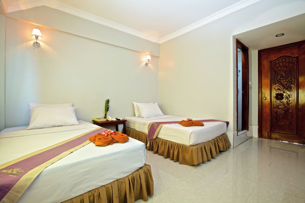 A hotel room with two beds, each with a white mattress, brown bed skirts, and orange towels. There are wall-mounted lights and a wooden door with a floral design.