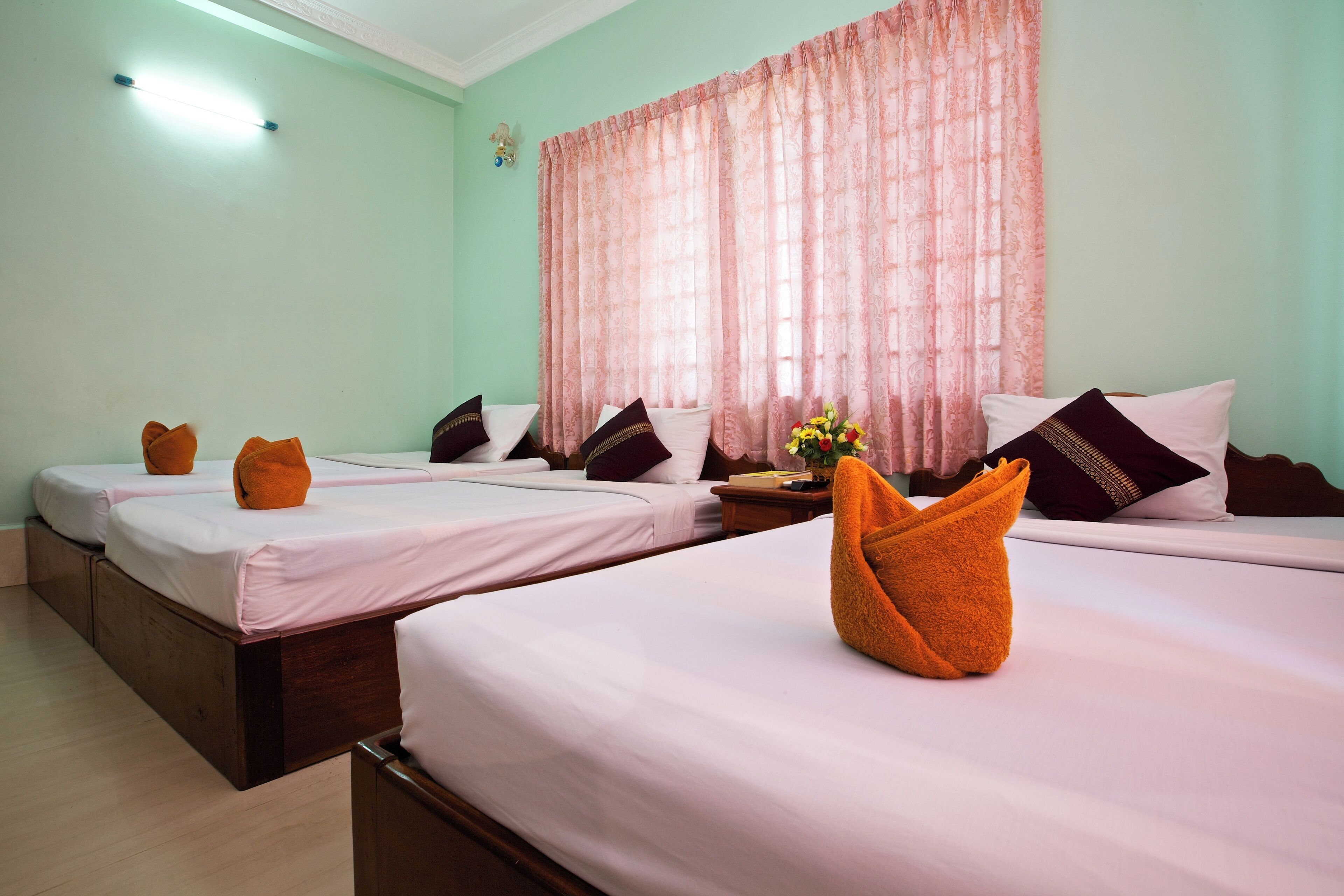 A hotel room with three beds, each with white linens and orange towels. There is a large window with pink curtains, and a small table with a vase of flowers.