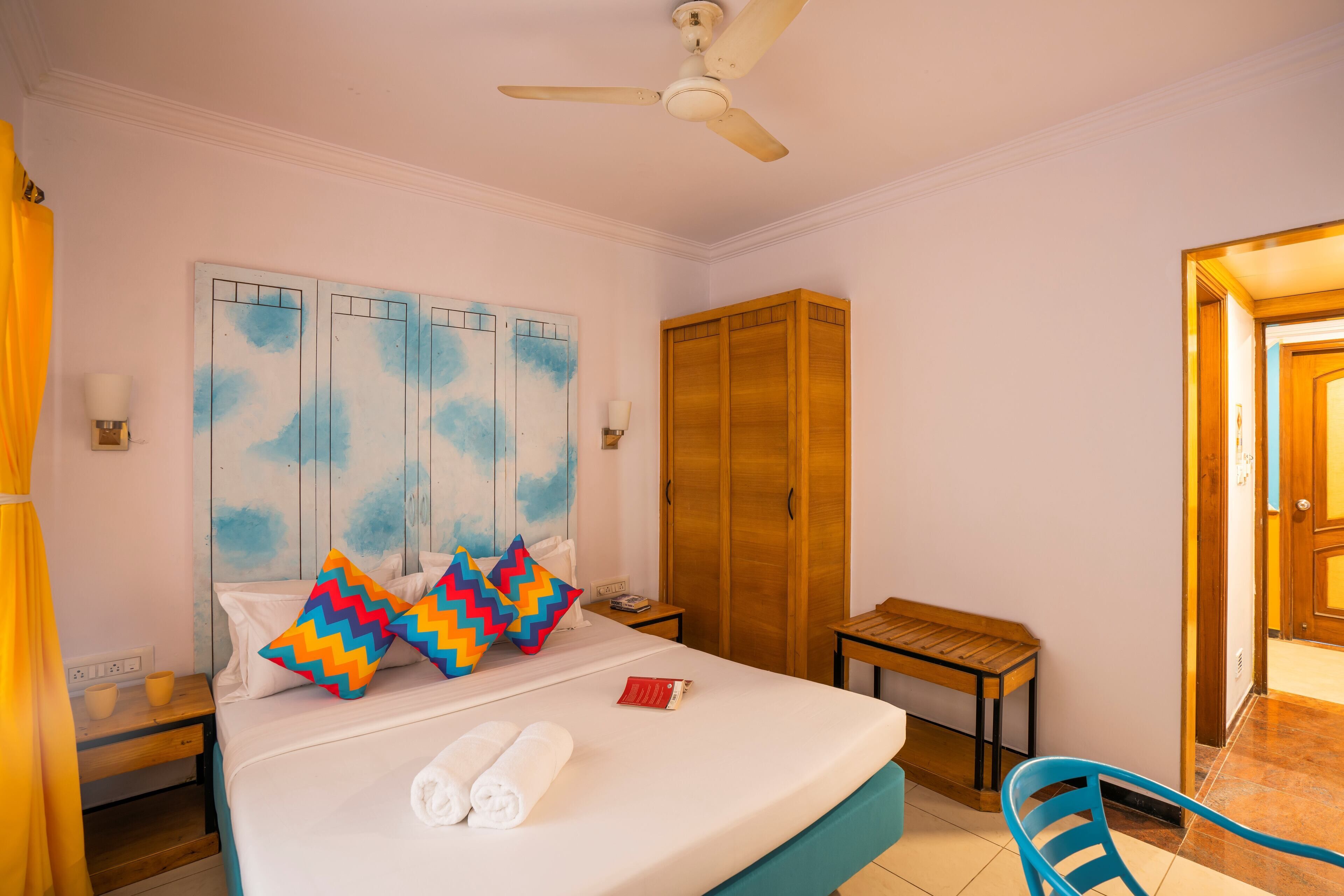 Une chambre d’hôtel comprenant un lit, une armoire en bois, une table de chevet avec une lampe, une petite table avec une chaise et un ventilateur de plafond.