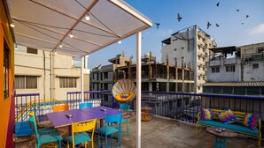 A rooftop terrace with colorful seating, a fan, and a view of surrounding buildings. - goSTOPS Bengaluru Gandhi Nagar Majestic. (Bengaluru)