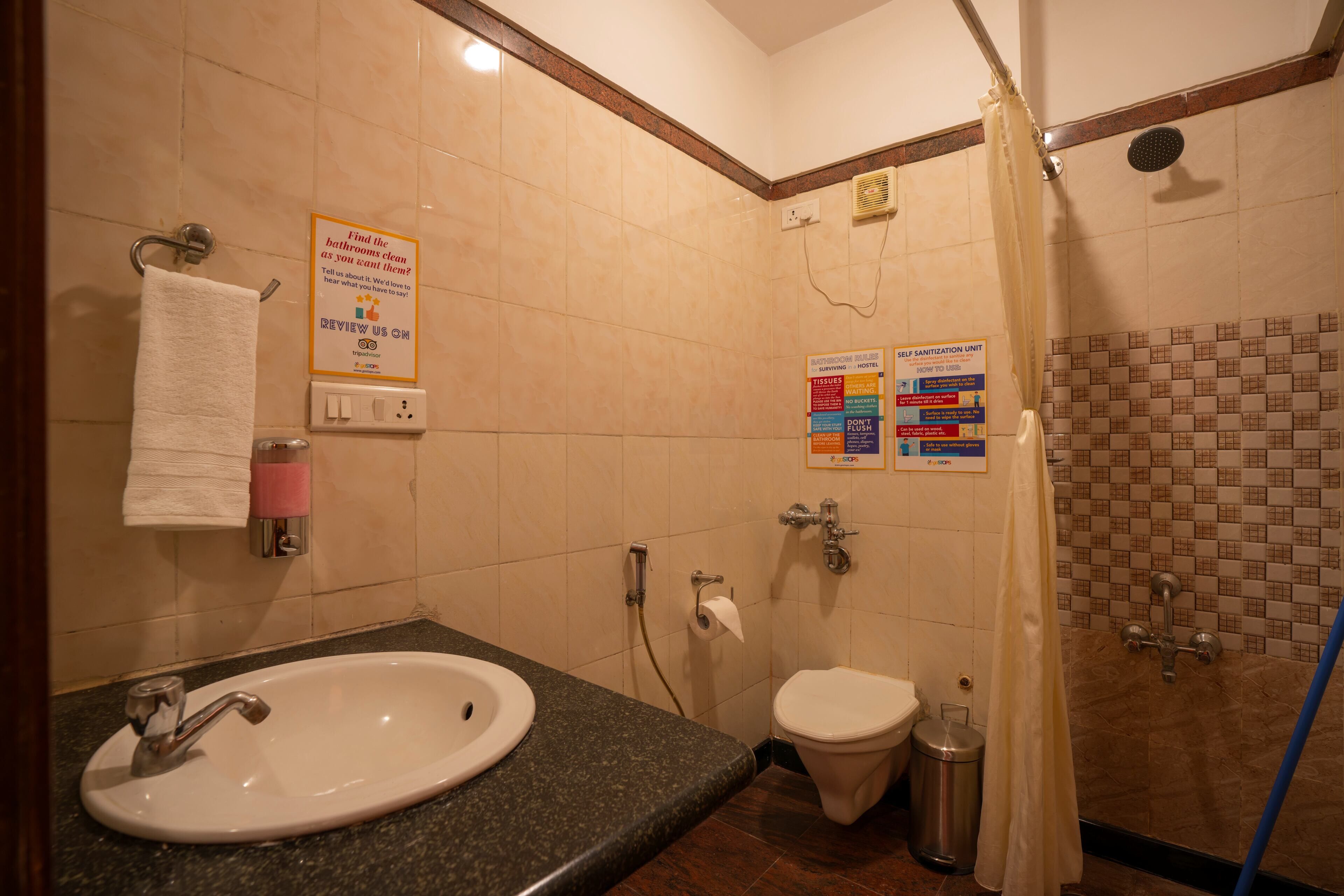 Un baño con lavamanos, inodoro, ducha y secamanos. Hay carteles en la pared con normas del baño y una unidad de auto-higienización.