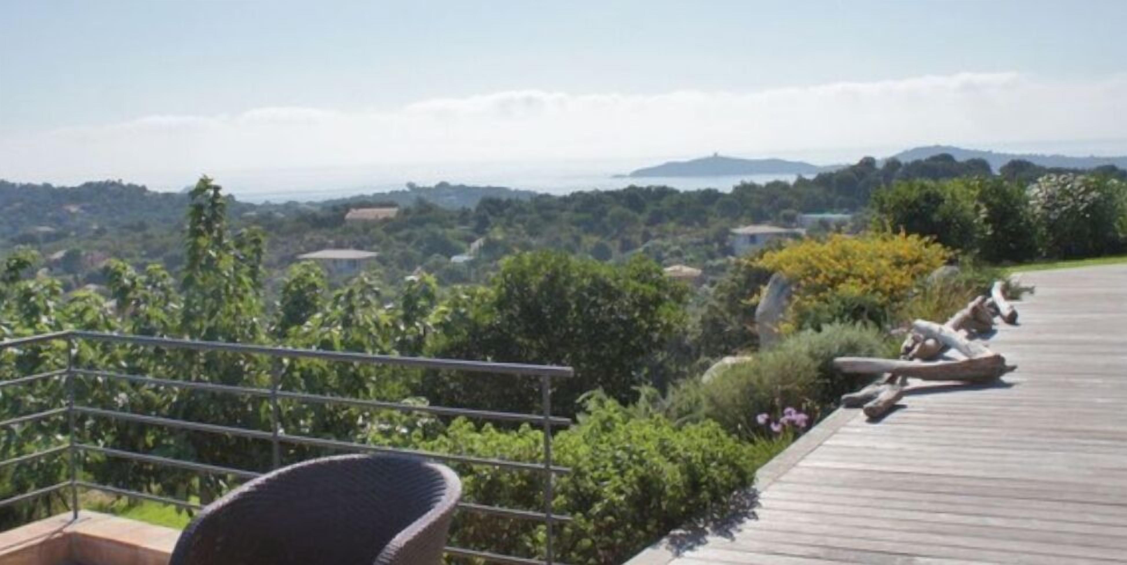 Un balcon avec un fauteuil en osier, une terrasse en bois et une vue sur un paysage verdoyant, avec des arbres et des collines à l’horizon.