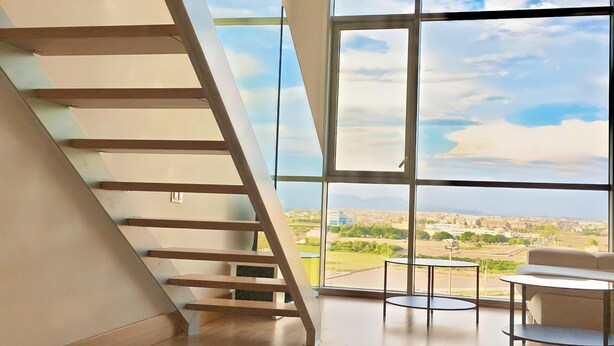 A spacious room with a wooden staircase, large windows offering a view of the landscape, and a seating area with a table.