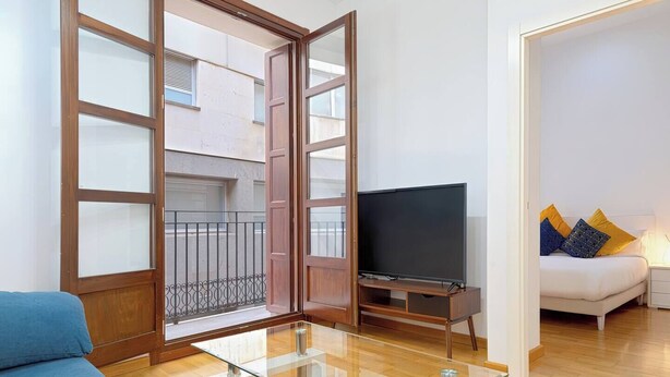 Un salon avec une table basse en verre, un canapé, un téléviseur posé sur un meuble en bois et un balcon doté d’une rambarde en métal.