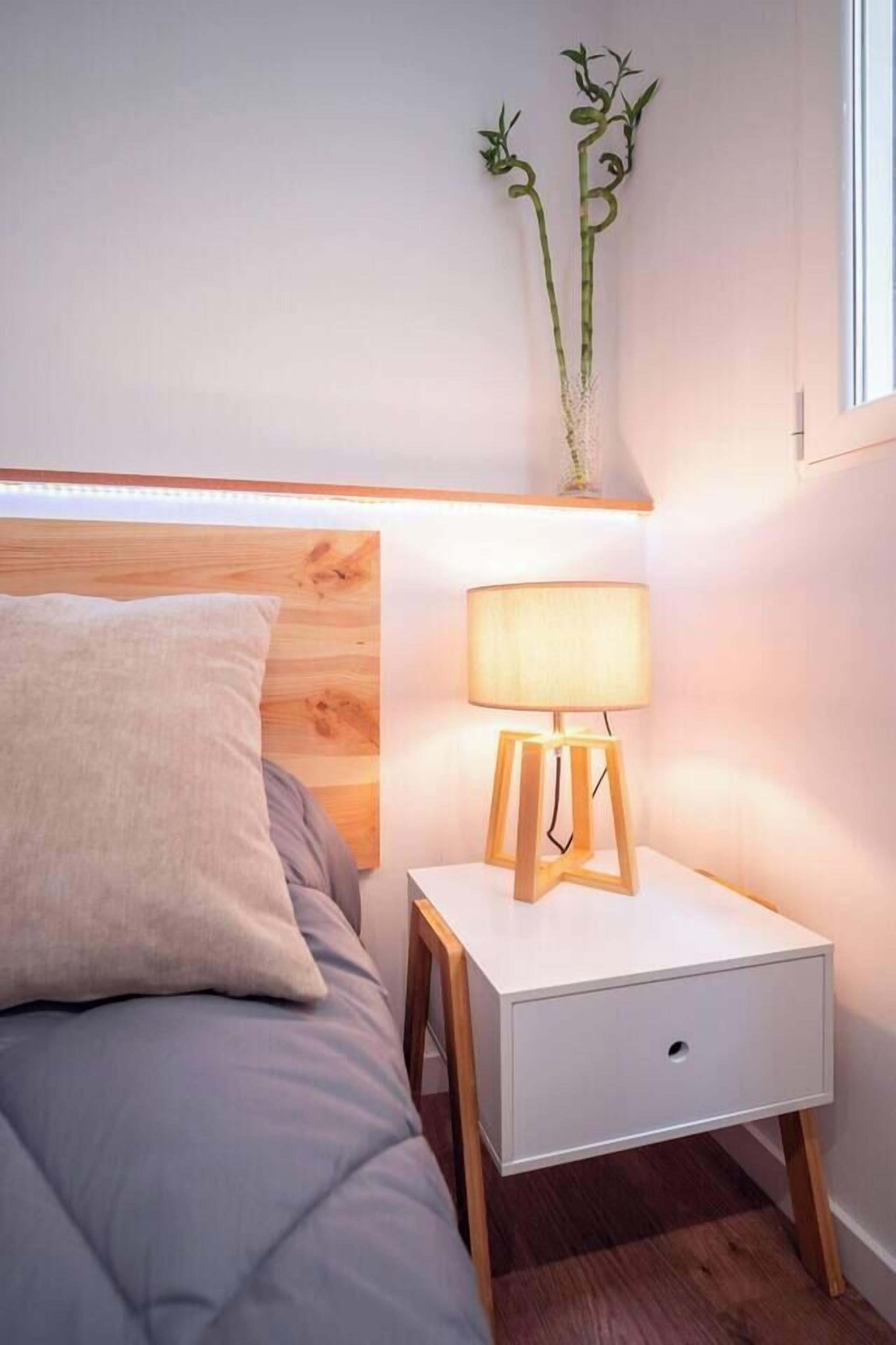 Een net opgemaakt bed met een kussen, een nachtkastje met een lamp en een aan de muur bevestigde plant.