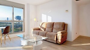 En moderne stue med en beige sofa, et glastæppe og udsigt over byen gennem store vinduer.